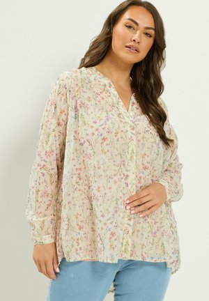 Kvinde med langt brunt hår iført en gennemsigtig cremefarvet blomsterknap-bluse og lyseblå jeans, poserer mod en ensfarvet baggrund.