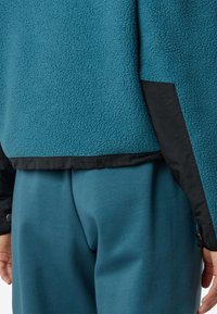 Giacca in fleece turchese con una finitura testurizzata e accenti neri, abbinata a pantaloni della tuta turchesi coordinati, che mostrano un look rilassato e stratificato.