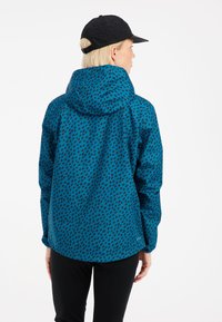 Mėlynas raštuotas džemperis su gobtuvu, turintis tekstūruotą juodų formų dizainą. Juoda kepurė, dėvima su tamsiomis kelnėmis.