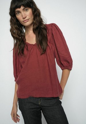 Žena s dlhými vlnitými hnedými vlasmi, oblečená v voľnej blúzke odtieňa hrdzavohnedej s nariaseným výstrihom a nadýchanými tromištvrťovými rukávmi, doplnená tmavými džínsami.