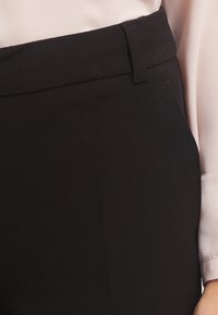 Detailní záběr osoby v černých kalhotách s vysokým pasem a v dlouhém svetru ve světle růžové barvě, na kterém jsou vidět pas a rukáv.