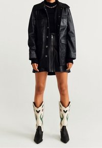 Chaqueta de cuero negra con bolsillos frontales, combinada con una minifalda negra. Botas de vaquero detalladas en crema con acentos florales en verde y rojo.