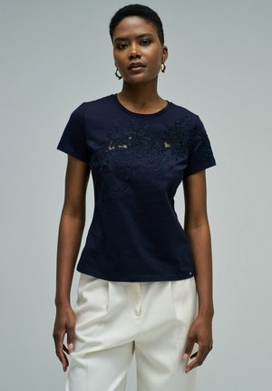 Femme portant une chemise bleu marine à manches courtes avec broderies florales et un pantalon blanc, debout devant un fond gris uni.