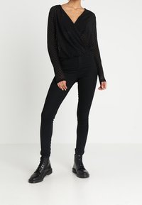 Camicetta nera trasparente a maniche lunghe con stampa a pois, scollatura a V profonda, design avvolgente, abbinata a pantaloni neri aderenti e stivaletti.