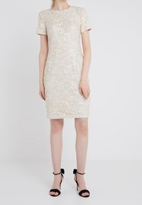 Žena v béžových krajkových šatech do kolen s krátkými rukávy a černých vysokých sandálech na podpatku s kotníkovými pásky a mašlemi.