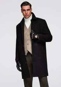 Mann trägt einen schwarzen Mantel, beige Weste, weißen Rollkragenpullover, braune Hose und schwarze Lederhandschuhe vor einem schlichten hellen Hintergrund.