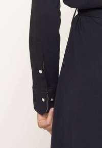 Chemise noire à manches longues avec des poignets boutonnés, présentant une texture lisse et un léger éclat. Coutures visibles le long des coutures.
