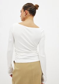Pitkähihainen, rib하셔하white top, jossa on leveä kaula-aukko. Siinä on tiukka siluetti ja rypytetty yksityiskohta takana, yhdistettynä vaaleanruskeaan hameeseen.