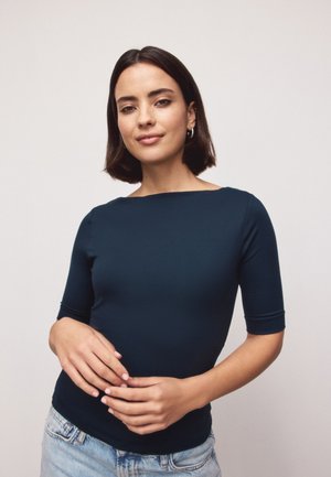 Junge Frau mit kurzen dunklen Haaren, die ein marineblaues, eng anliegendes Oberteil und hellblaue Jeans trägt, steht mit sanft verschränkten Händen.