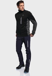 Schwarze und graue Zip-Jacke mit hohem Kragen, zwei seitlichen Taschen und reflektierenden Akzenten, kombiniert mit marineblauen Hosen mit Reißverschlusstaschen und schwarzen Schuhen.