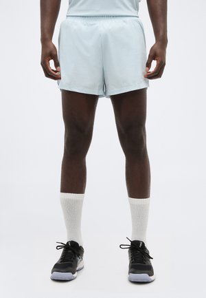 Άνδρας με ανοιχτό μπλε αθλητικό σορτς, λευκές κάλτσες μέχρι τον αστράγαλο και μαύρα αθλητικά παπούτσια, στέκεται με τα χέρια χαλαρά στο πλάι σε λευκό φόντο.