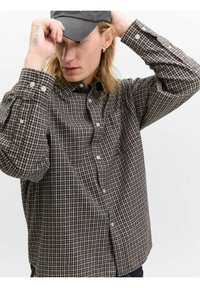 Ung person med langt hår, der justerer en sort kasket, iført guld hoop ørering og ternet skjorte med knapper samt en teksttattoo på hånden.