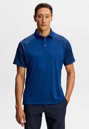 Uomo che indossa una polo blu con maniche corte a motivi, in piedi con una mano in tasca davanti a uno sfondo semplice.