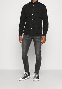 Zwart overhemd met knopen en twee borstzakken, gecombineerd met strakke grijze jeans en zwarte sneakers. Glad materiaal met een klassieke kraag.