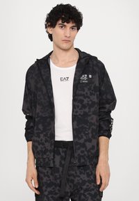 Black and gray patterned jacket with a hood, featuring a front zipper and logo accents. Worn over a white t-shirt and paired with dark pants.