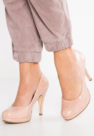 Jasnoróżowe lakierowane szpilki z okrągłym noskiem, platformą z przodu i smukłym obcasem. Noszone z podwiniętymi jasnoszarymi spodniami.