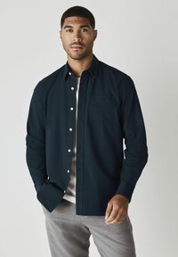 Camicia button-up di cotone blu scuro, con tasca, bottoni bianchi e maniche lunghe. Indossata sopra una maglietta chiara.