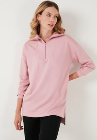 Pullover rosa chiaro con collo zip, maniche lunghe e fessure laterali, realizzato in un morbido tessuto e caratterizzato da una texture liscia.
