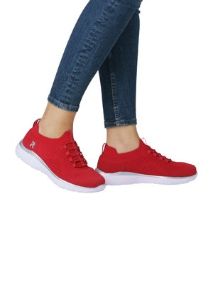 Rote Sportschuhe aus gestricktem Stoff mit strukturierten Akzenten, ausgestattet mit einer weißen Sohle und elastischen Schnürsenkeln, getragen von einer Person in blauen Jeans.