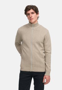 Suéter beige con cremallera y cuello alto, que presenta un patrón en chevrón texturizado y puños y dobladillo acanalados, combinado con pantalones oscuros.
