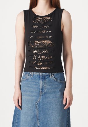 Mujer con camiseta negra sin mangas con cortes horizontales que revelan encaje negro debajo, combinada con falda de mezclilla azul de cintura alta.