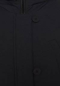 Svart kappa med dragkedjestängning, två stora knappar och en minimalistisk design. Slät textur med en elegant, modern look.