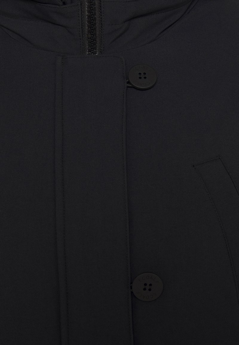 Svart kappa med dragkedjestängning, två stora knappar och en minimalistisk design. Slät textur med en elegant, modern look.