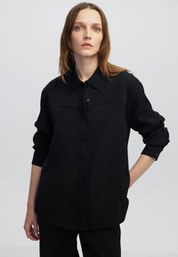 Camicia nera a bottoni con colletto, maniche lunghe e tessuto testurizzato. Due tasche frontali delineate da un pannello a contrasto.