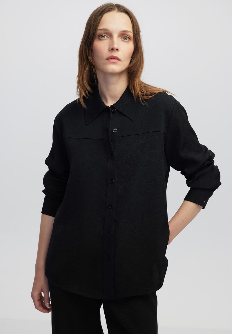 Camicia nera a bottoni con colletto, maniche lunghe e tessuto testurizzato. Due tasche frontali delineate da un pannello a contrasto.