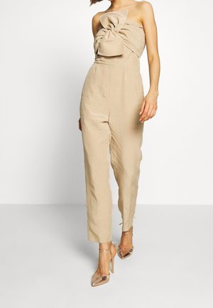 Combinaison en lin beige avec un grand nœud au niveau de la poitrine, rayures verticales orange clair, coupe ajustée. Associée à des talons transparents.