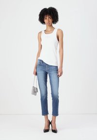 Weißes ärmelloses Tanktop, hellblaue enganliegende Jeans und schwarze High Heels. Das Model hält eine kleine silberne Handtasche.