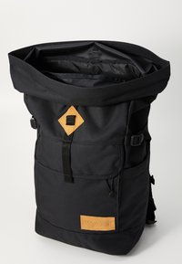 Musthave rullitask, mis on valmistatud kangast, musta värvi, nahkdetailiga, reguleeritavate õlapaelte ja välistaskutega.