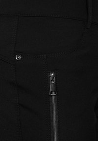 Černé kalhoty vyrobené z pružné látky mají boční zipovou kapsu a stříbrné kovové doplňky, včetně cvokového tlačítka a kontrastního šití.