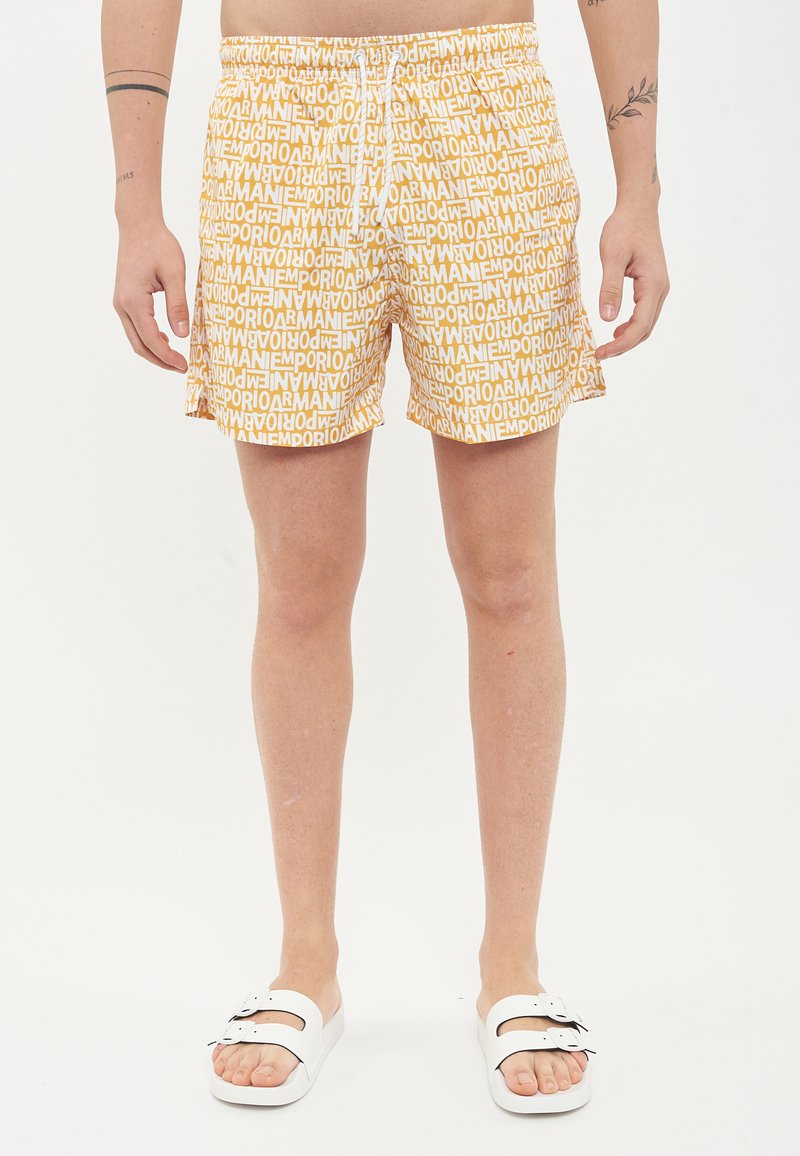 Pantaloni da nuoto corti con cintura elastica, caratterizzati da una stampa all-over del logo in bianco su uno sfondo giallo. Indossati con sandali scivolati bianchi.