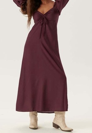 Mujer con un vestido maxi morado oscuro de manga larga con un detalle de lazo frontal, combinado con botas beige claras hasta media pantorrilla, de pie con los brazos levantados.
