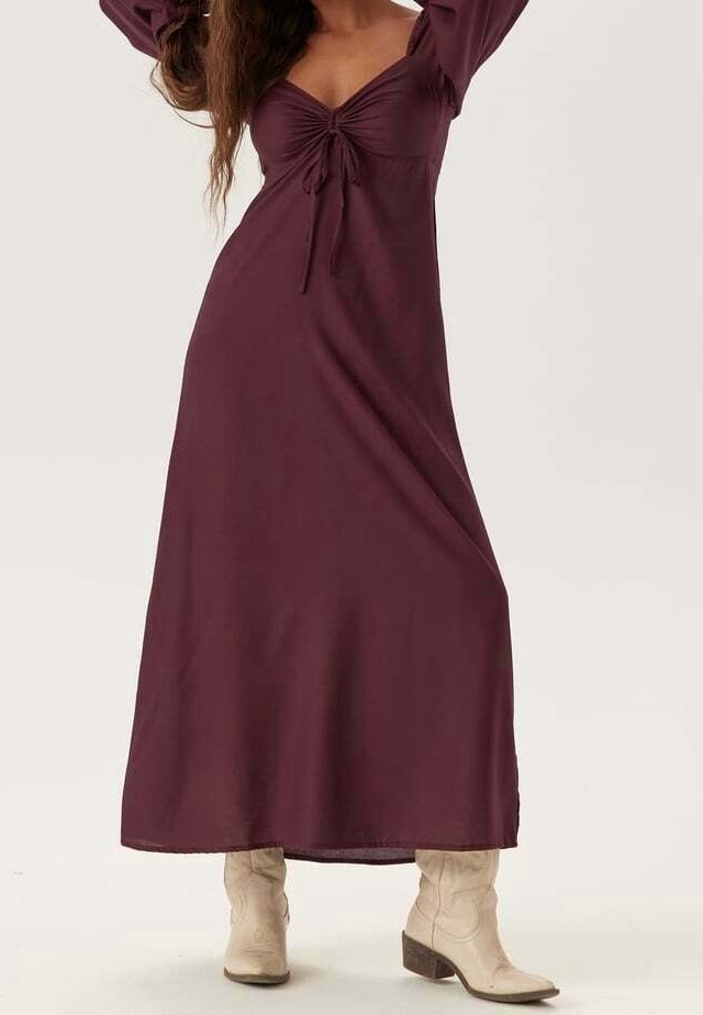 Femme portant une robe longue à manches longues de couleur violet profond avec un détail noué à l'avant, assortie à des bottes de couleur beige clair jusqu'au milieu du mollet, debout avec les bras levés.