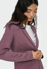 Lilla skreddersydd blazer, myk stoff, notcheskåpe, enkel knappelukking, sidelommer, båret over en hvit skjorte, slank silhuett.