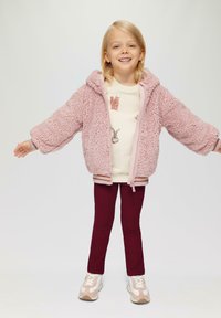 Roze donzige jas met capuchon, crèmekleurig shirt met dierenprints, bordeauxrode legging en lichte sneakers met subtiele accenten.