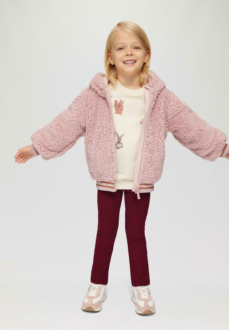 Roze donzige jas met capuchon, crèmekleurig shirt met dierenprints, bordeauxrode legging en lichte sneakers met subtiele accenten.