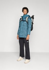 Teal dunjakke med horisontale sømmer, frontglidelås og hette. Svarte cargo-bukser med lommer, kremfargede joggesko og en grå ryggsekk.