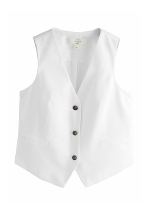 Donna con gilet bianco senza maniche e pantaloni bianchi a gamba larga in piedi contro una parete beige con grata decorativa in ferro della finestra.