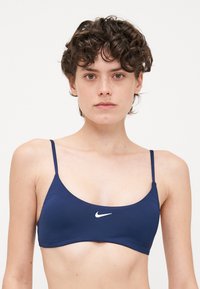 Temno modro bikini zgornji del z tankimi nastavljivimi trakovi, gladko teksturo in majhnim belim logotipom Nike na sredini sprednje strani. Preprost dizajn.
