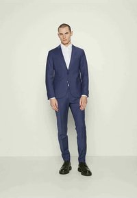 Homem em pé, vestido com um fato azul marinho justo, camisa branca e sapatos de vestir pretos, contra um fundo bege liso.