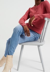 Červený svetr s texturou pleteniny, kulatým výstřihem a zapínáním na knoflíky, v kombinaci s modrými úzkými džíny a béžovými kotníkovými botami, sedící na šedé židli.