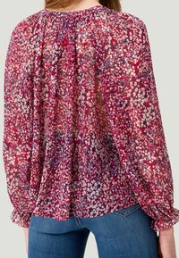 Gennemsigtig, let bluse med et blomsterprint i røde, pink og blå nuancer. Den har en sammensnøring ved halsen og elastiske manchetter.