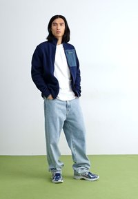 Navy fleece jacket with a teal zippered pocket, white t-shirt, light blue loose jeans, and gray-blue athletic shoes. Simple design, casual fit.