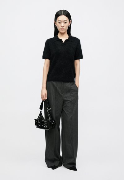 Sort, blødt poloshirt med korte ærmer, kombineret med brede grå bukser med striber. Modellen holder en lille sort taske med sølvdetaljer.