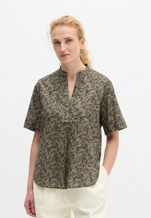 Femme aux cheveux blonds bouclés portant une chemise florale olive à manches courtes et un pantalon blanc, debout devant un fond blanc uni.