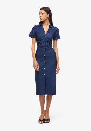 Naine seisab seljas tihedalt istuv keskmise pikkusega tume sinine nööpidega kleit lühikeste varrukatega ja mustade avatud varbaga kontsakingadega, vaadates vasakule.