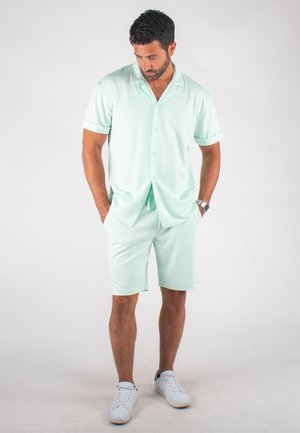Chemise à manches courtes vert menthe clair avec un col, associée à un short assorti. Portée avec des baskets blanches, présentant une texture lisse.
